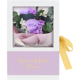 A floral gift box for Mom featuring a bouquet of purple and white flowers with green eucalyptus leaves inside a clear window, wrapped in pink paper with a white ribbon. The box has a lavender lower section with gold script reading 'Beautiful Mom' and a yellow ribbon tied to the side.